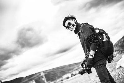 Low angle view of man with camera wearing skull mask while standing against sky