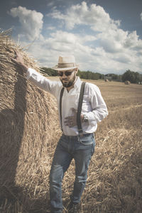 Man wearing hat standing by hay bale on land against sky