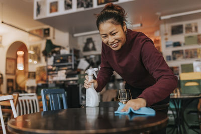 Happy female coffee shop owner with spray bottle and rag cleaning table in cafe