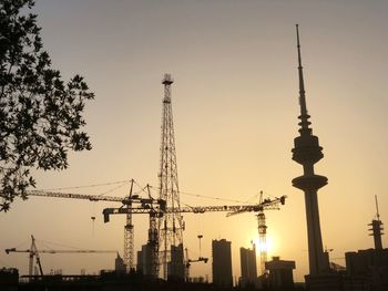 Silhouette cranes by buildings against sky during sunset