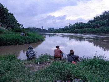 Rear view of men sitting on riverbank against sky