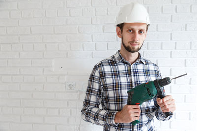 Portrait of man holding drill standing against wall