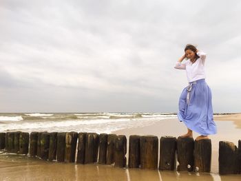 Low angle view of young woman standing on wooden posts in sea against sky