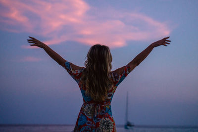 Rear view of woman with arms raised against sky during sunset