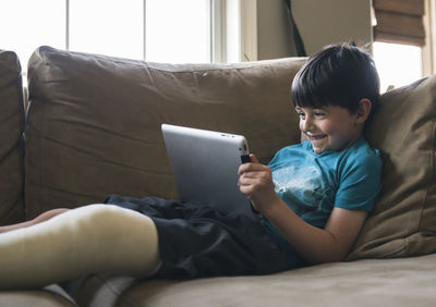 Smiling boy with fractured leg using tablet computer while lying on sofa at home