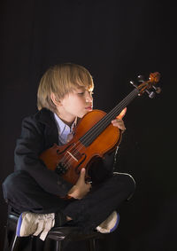 Boy holding violin while sitting against black background