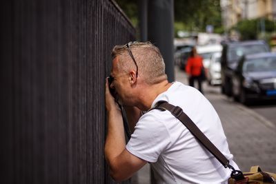 Man looking through fence while standing on sidewalk in city