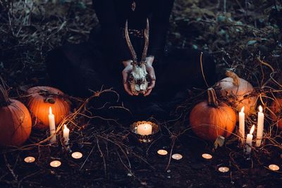 Low section of woman holding animal skull while sitting amidst pumpkins and burning candles during halloween