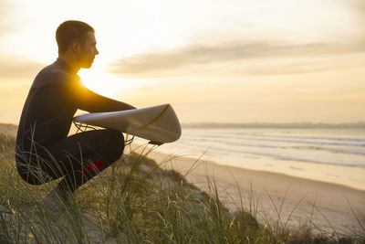 Side view of man sitting on beach against sky during sunset