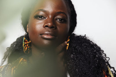 Portrait of young curvy woman wearing stylish african clothes looking at camera while standing in light room. she has unfocused plants in the foreground.