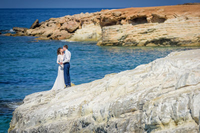 Newlywed couple kissing while standing on rock by sea