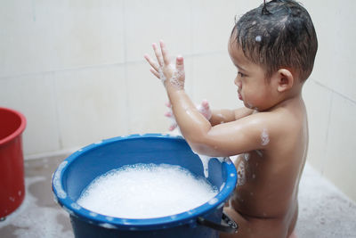 Shirtless boy playing with soap sud while sitting in bathroom