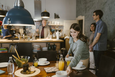 Smiling teenage girl placing bowls on dining table for dinner with family at home