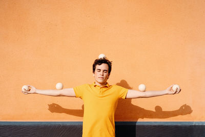 Tranquil immobile young male in bright yellow shirt with juggling balls on head and outstretched arms standing with eyes closed against orange wall