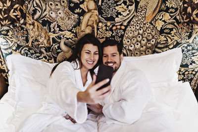 Happy mid adult woman and man taking selfie on bed in hotel room