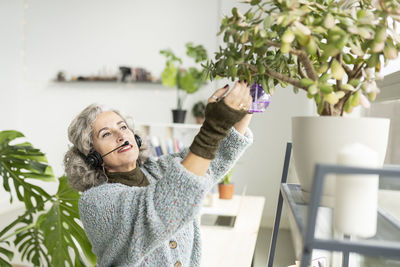 Senior businesswoman wearing headset spraying water on potted plants at home