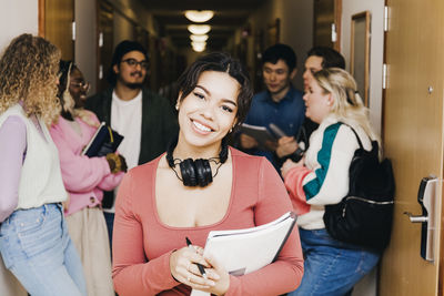 Portrait of smiling young woman with book while multiracial friends in background at corridor in college dorm