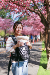 Portrait of woman with baby carriage standing by cherry blossom in park