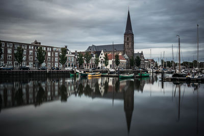 Sailboats moored on river by buildings against cloudy sky