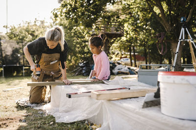 Daughter looking at mother using drill machine on wooden plank in yard