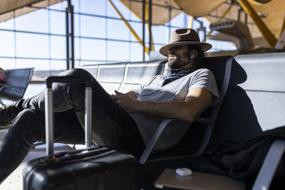 Adult bearded man in stylish outfit with hat sitting in a bench sleeping next to glass wall listening to music on wireless earphones while waiting for flight in airport lounge