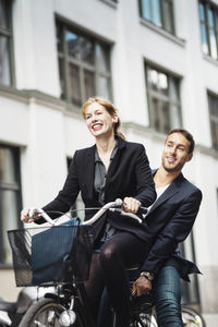 Happy businesswoman riding bicycle while colleague sitting on back seat outdoors
