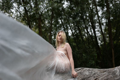 Portrait of naked woman wrapped in a polythene sitting on tree trunk in forest