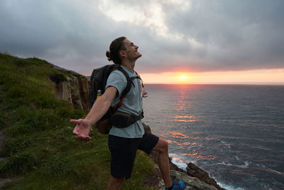 Side view of carefree male hiker standing at viewpoint on rock and enjoying freedom on background of sunset over sea