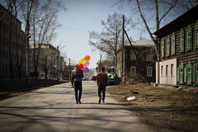 Rear view of man with balloons walking by friend on road in city