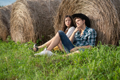 A young man with a straw in his mouth sits near a roll of hay and looks into the distance. 