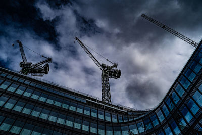 Low angle view of cranes on building against cloudy sky