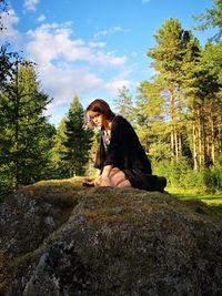 Girl sitting on rock at forest against sky