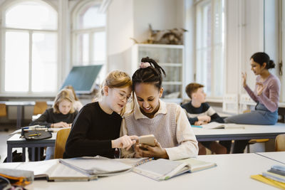 Curious schoolgirls sharing smart phone while sitting at desk in classroom