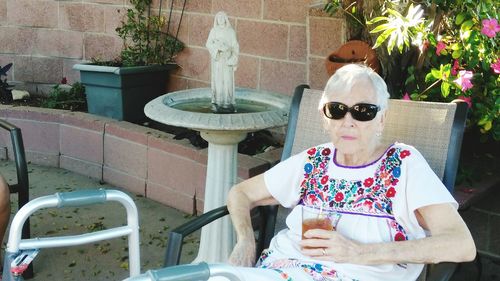 Senior woman holding drink while sitting on deck chair at back yard