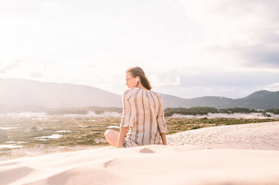 Rear view of woman sitting at sandy beach against sky