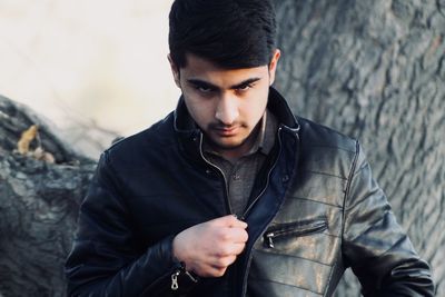 Portrait of young man wearing leather jacket while standing against tree