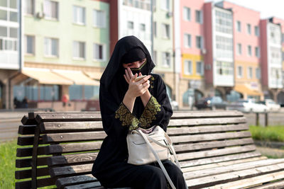 A muslim woman is resting on a bench in the park, tidying herself up looking in a mirror.