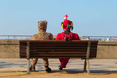 Full length rear view of friends in costumes sitting on bench against clear blue sky