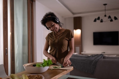 Cheerful mixed race female in headphones chopping cucumber