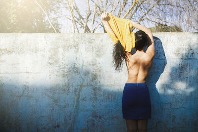 Rear view of shirtless woman removing top while standing against wall