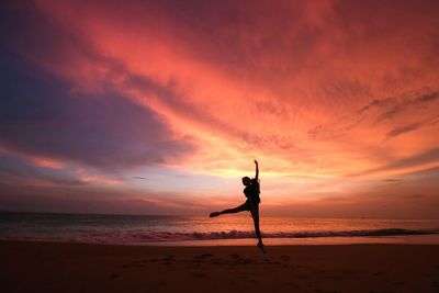 Silhouette person jumping on beach against multi colored cloudy sky
