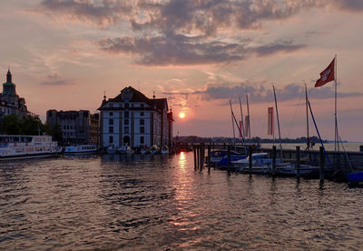 Sailboats moored in harbor by buildings against sky during sunset