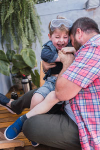 Unrecognizable bearded dad in checkered shirt playing with cheerful boy in safety glasses while sitting in daytime