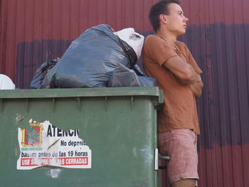 Side view of thoughtful young man standing with arms crossed by garbage bin
