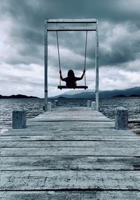 Rear view of woman sitting on swing over pier and sea against sky