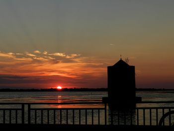 Silhouette building by sea against orange sky