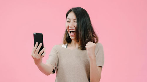 Young woman using phone while standing against pink background