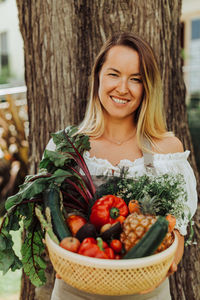 Young smiling woman with basket of vegetables standing against a tree