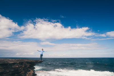 Man with arms outstretched standing on cliff by sea against blue sky