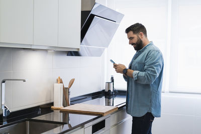 Man cooking crepes in the kitchen with a mobile phone in a denim shirt